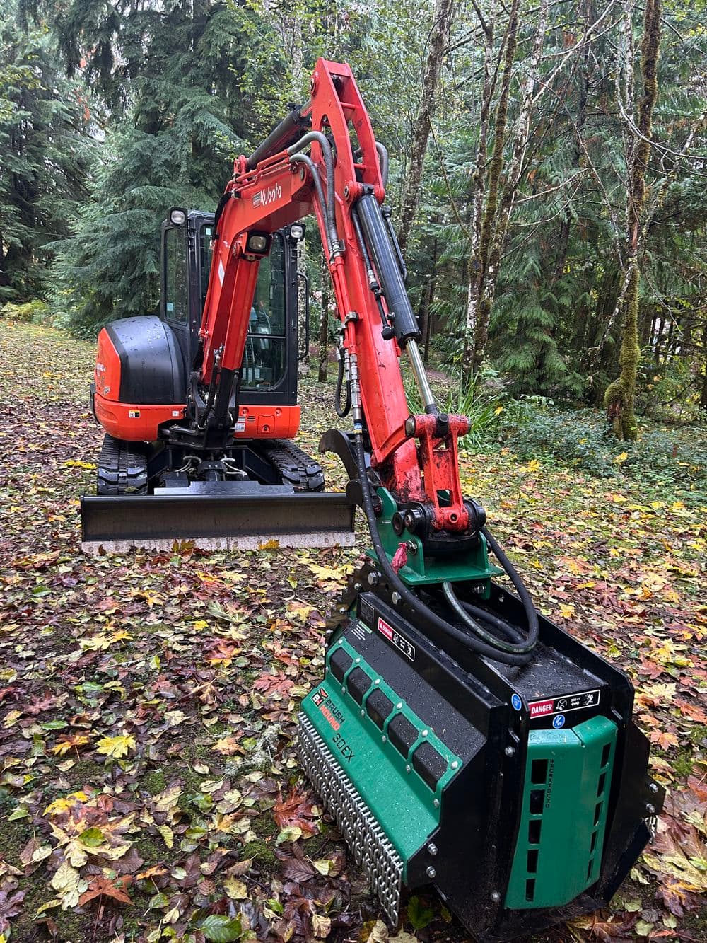 Excavator with brush attachment in forested area with fallen leaves on the ground.